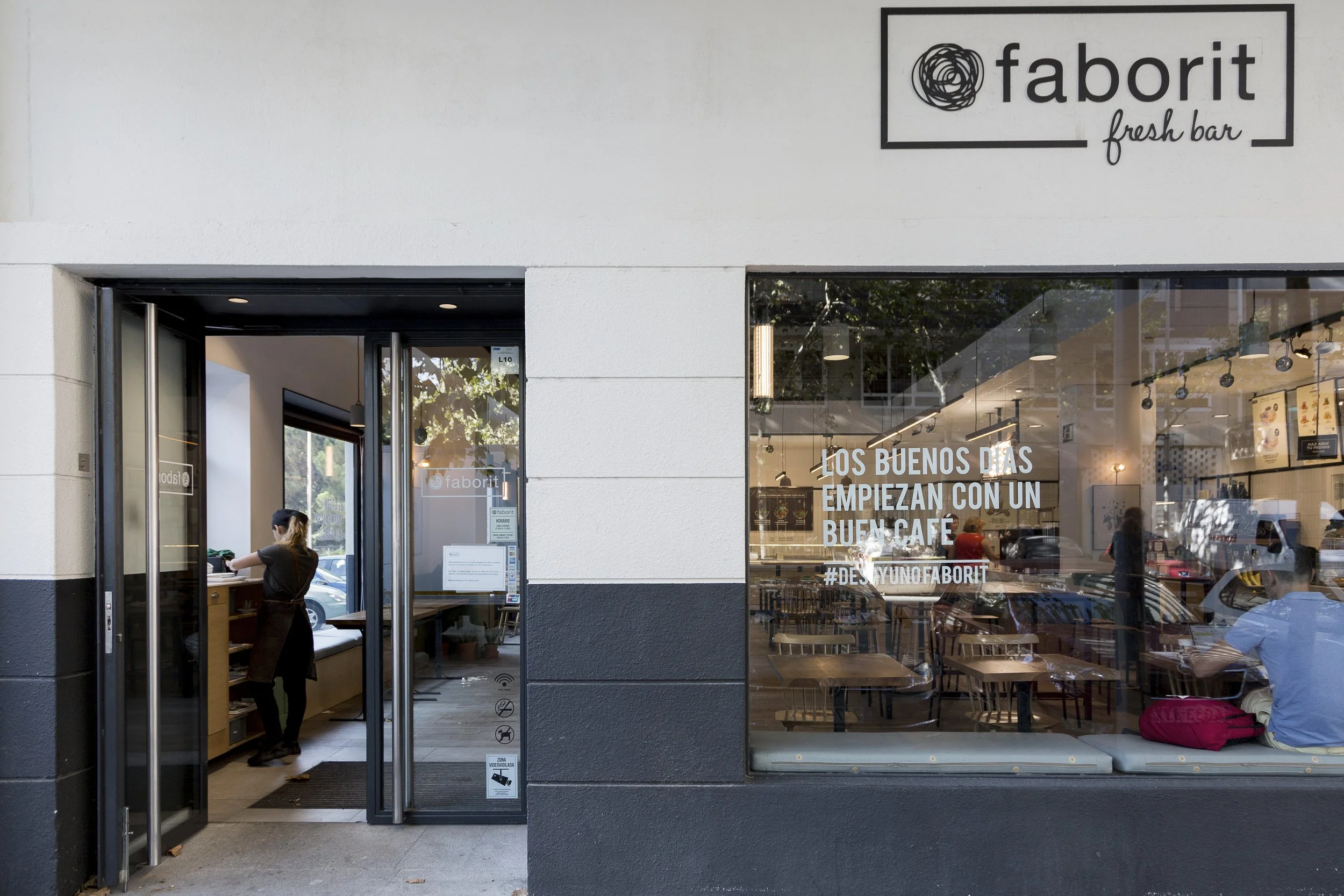 Entrada de cafetería con el letrero 'faborit', vista del interior con mesas y sillas, y una persona adentro preparando algo en la barra. Texto en la ventana dice 'Los buenos días empiezan con un buen café'. Diseño de interiorismo comercial.