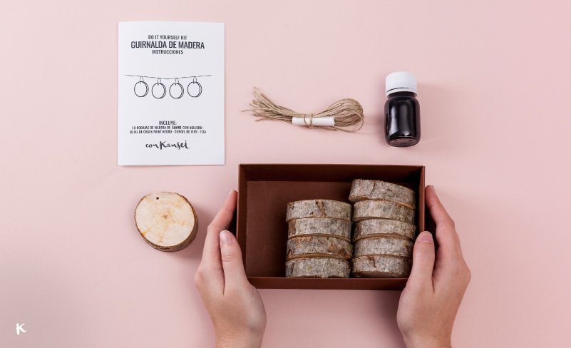 Un kit de bricolaje para hacer decoración con anillos de madera, incluyendo anillos de madera, cuerda de yute, pintura negra y instrucciones en papel, sobre fondo rosa