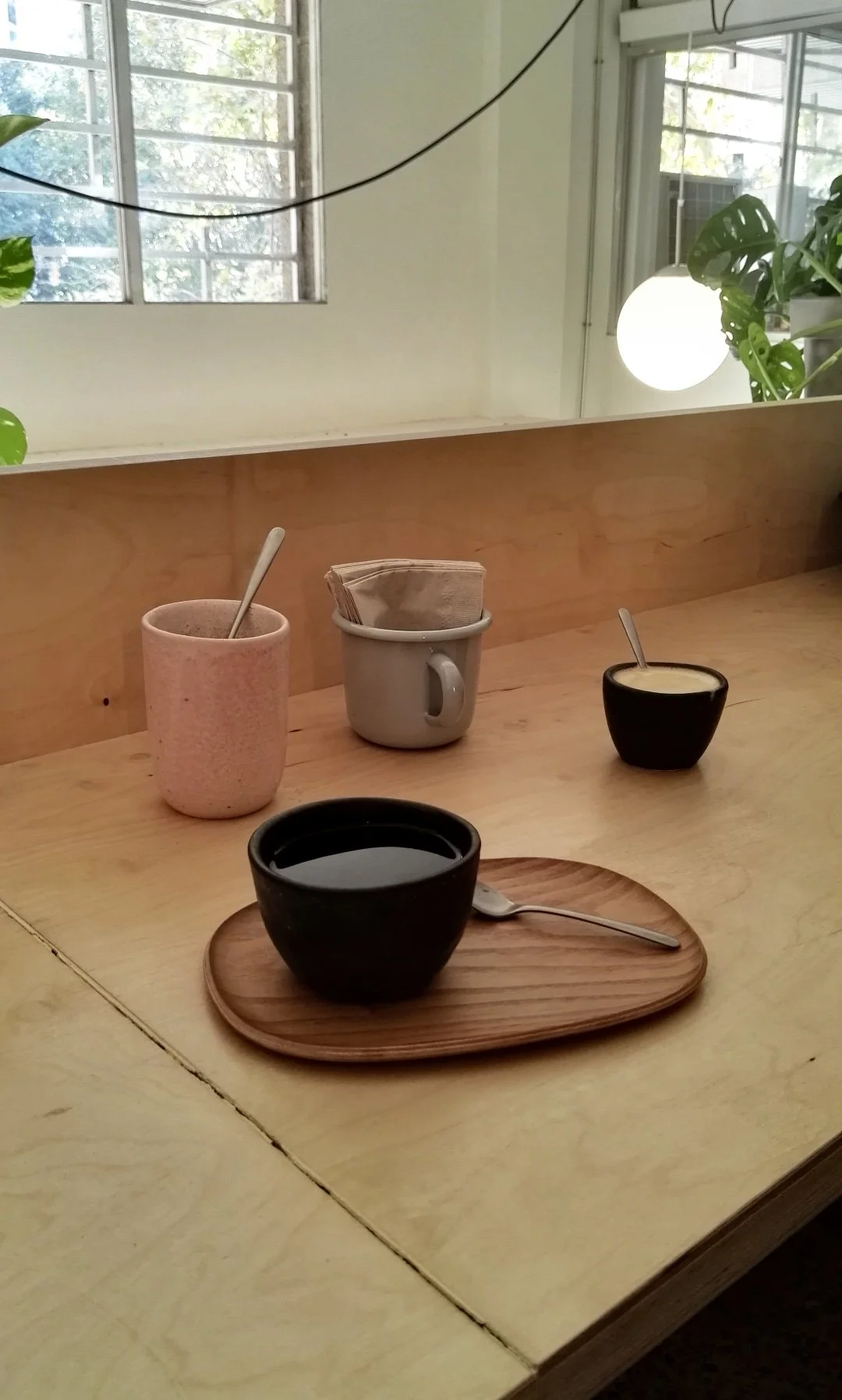 Cuatro tazas y un plato de madera con una cuchara en un mostrador de madera en un ambiente con plantas y ventanas grandes.