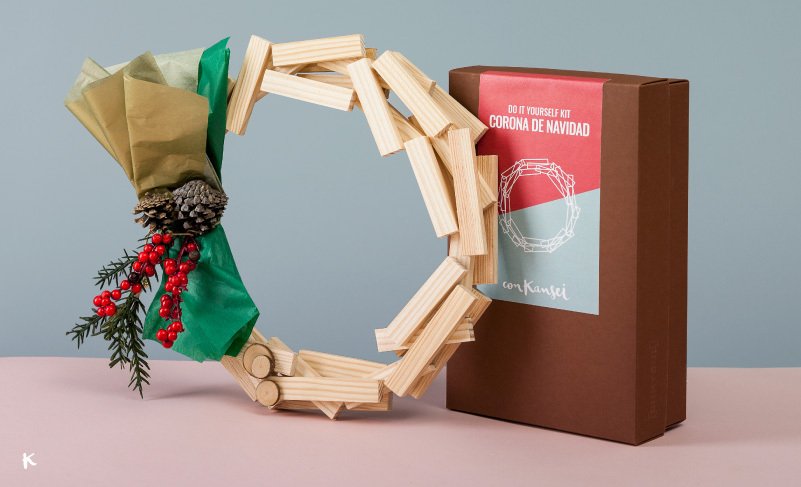 Corona de Navidad hecha con bloques de madera, decorada con hoja de acebo, piña y ramas, junto a una caja de regalos con kit de corona navideña DIY.
