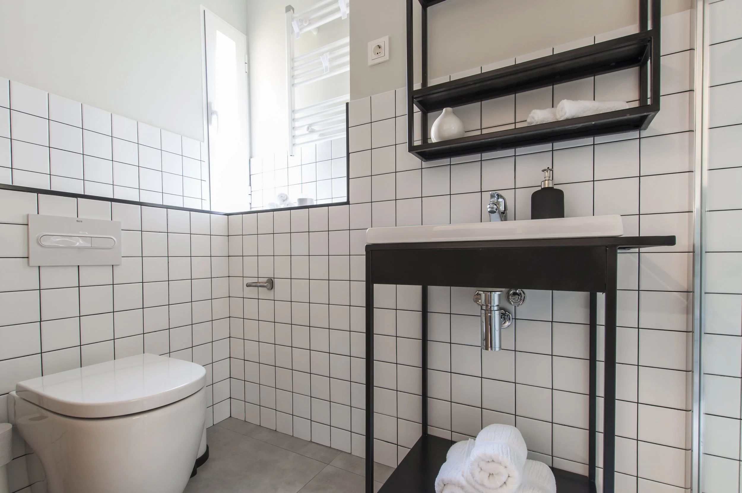 Baño moderno con azulejos blancos, lavamanos negro, toallas enrolladas y un inodoro.