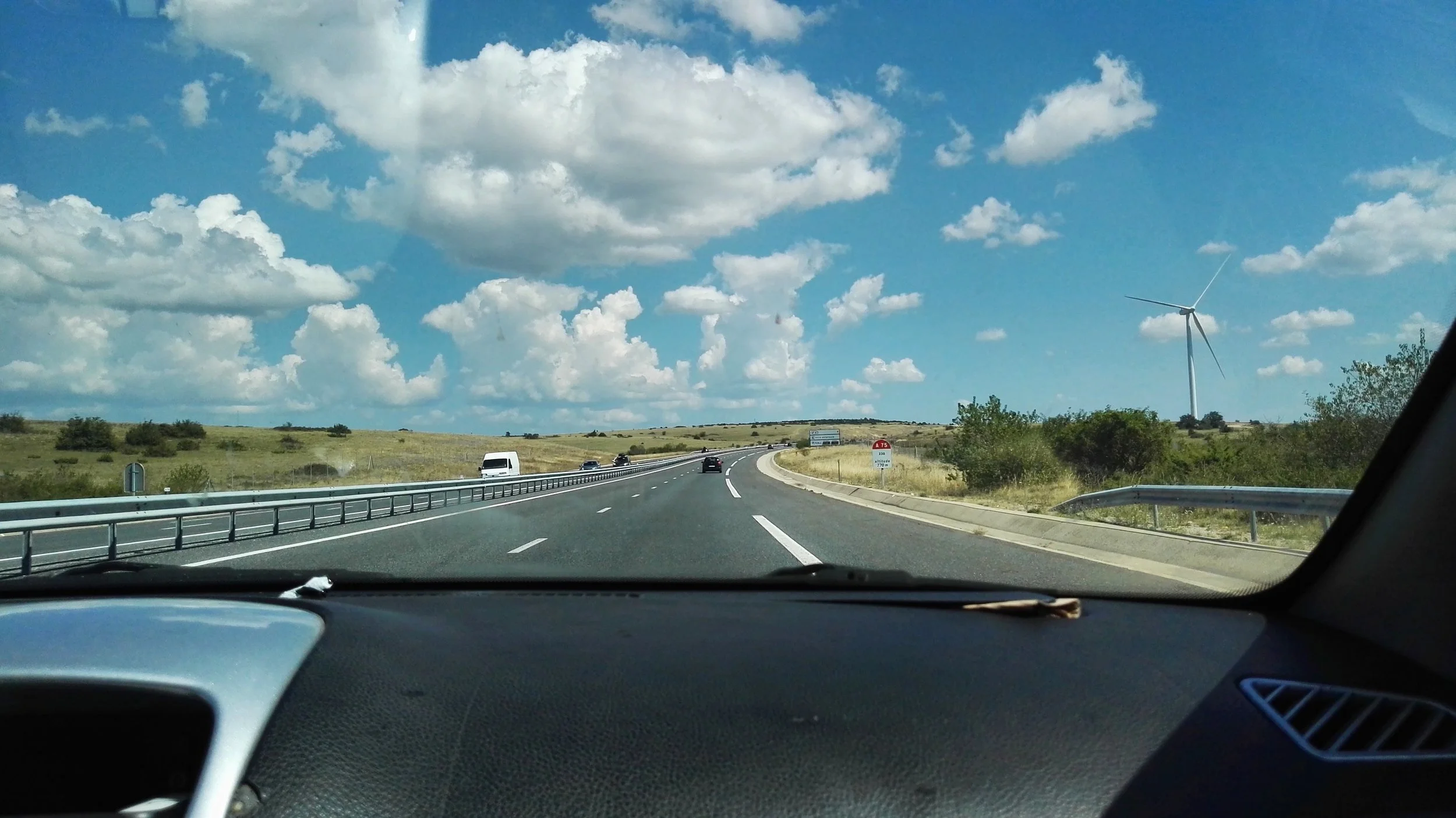 Vista de una autopista con pocos autos en un día soleado, rodeada de campos y un aerogenerador en la distancia. Roadtrip.