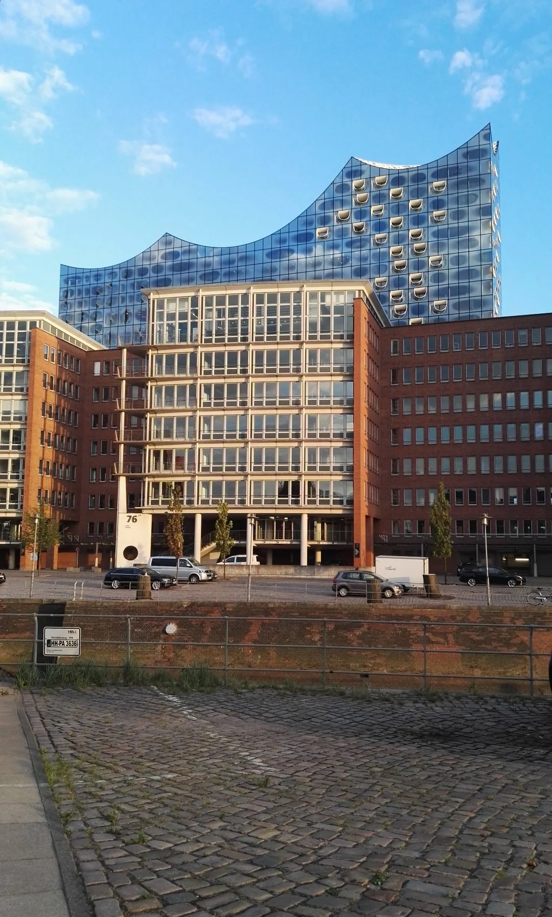 Edificio moderno con fachada de vidrios azules y forma ondulada en la parte superior, junto a edificios de ladrillo, en una ciudad bajo cielo despejado. Hamburg Philarmonic.