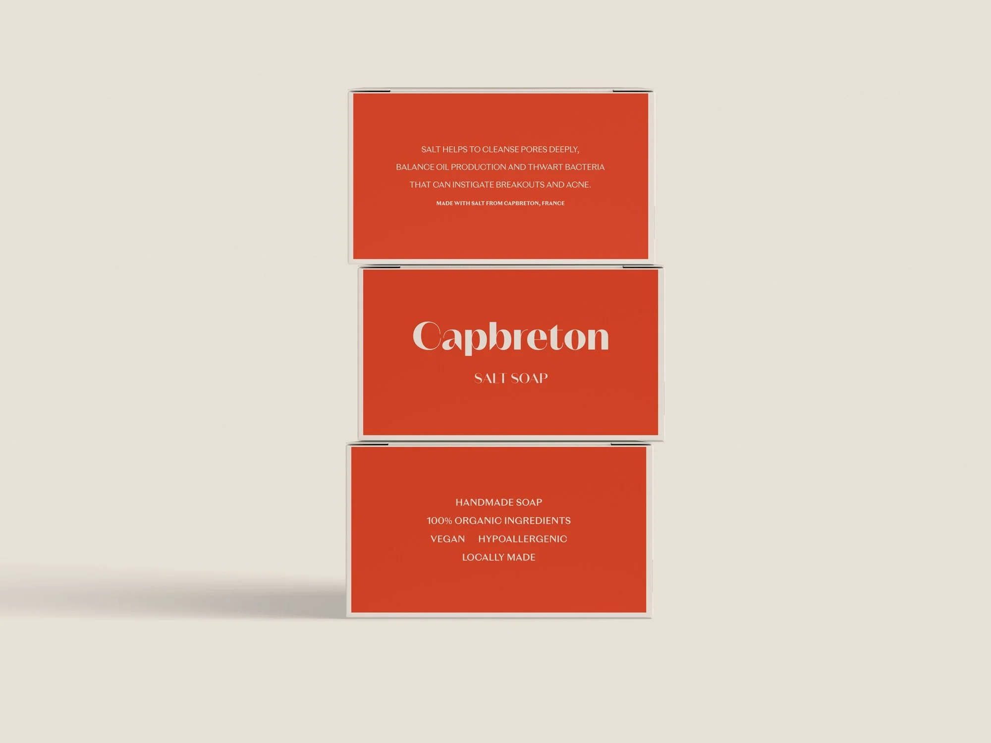 Tres cajas apiladas de jabón "Capbreton Salt Soap" en un fondo claro. Las cajas son de color rojo con texto blanco que describe los beneficios del jabón. Indican que el jabón es hecho a mano, con ingredientes 100% orgánicos, vegano, hipoalergénico y