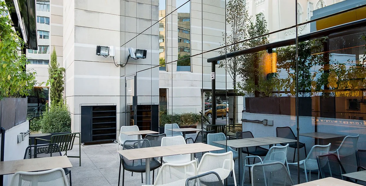 Terraza exterior de un café con mesas y sillas. Al fondo se ven edificios reflejados en las ventanas de vidrio y hay plantas decorativas. Diseño de interiorismo comercial. Retail Design. Foto de Eva Carasol.
