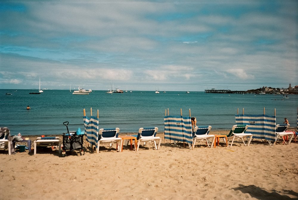 Playa con sillas y sombrillas, agua azul, barcos y un muelle en el fondo.
