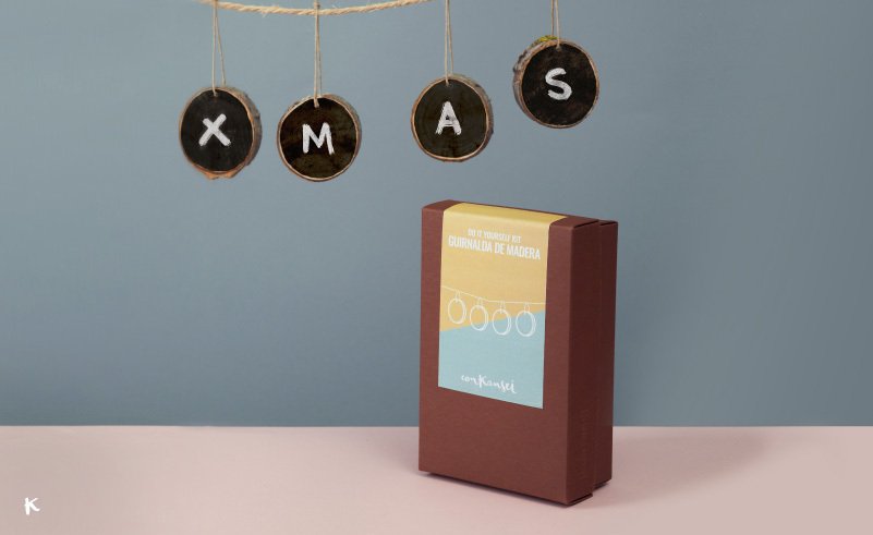 Paquete de una cuerda de madera en una mesa, con letras en círculos colgados que forman la palabra 'XMAS', y caja de madera con etiqueta en el fondo.