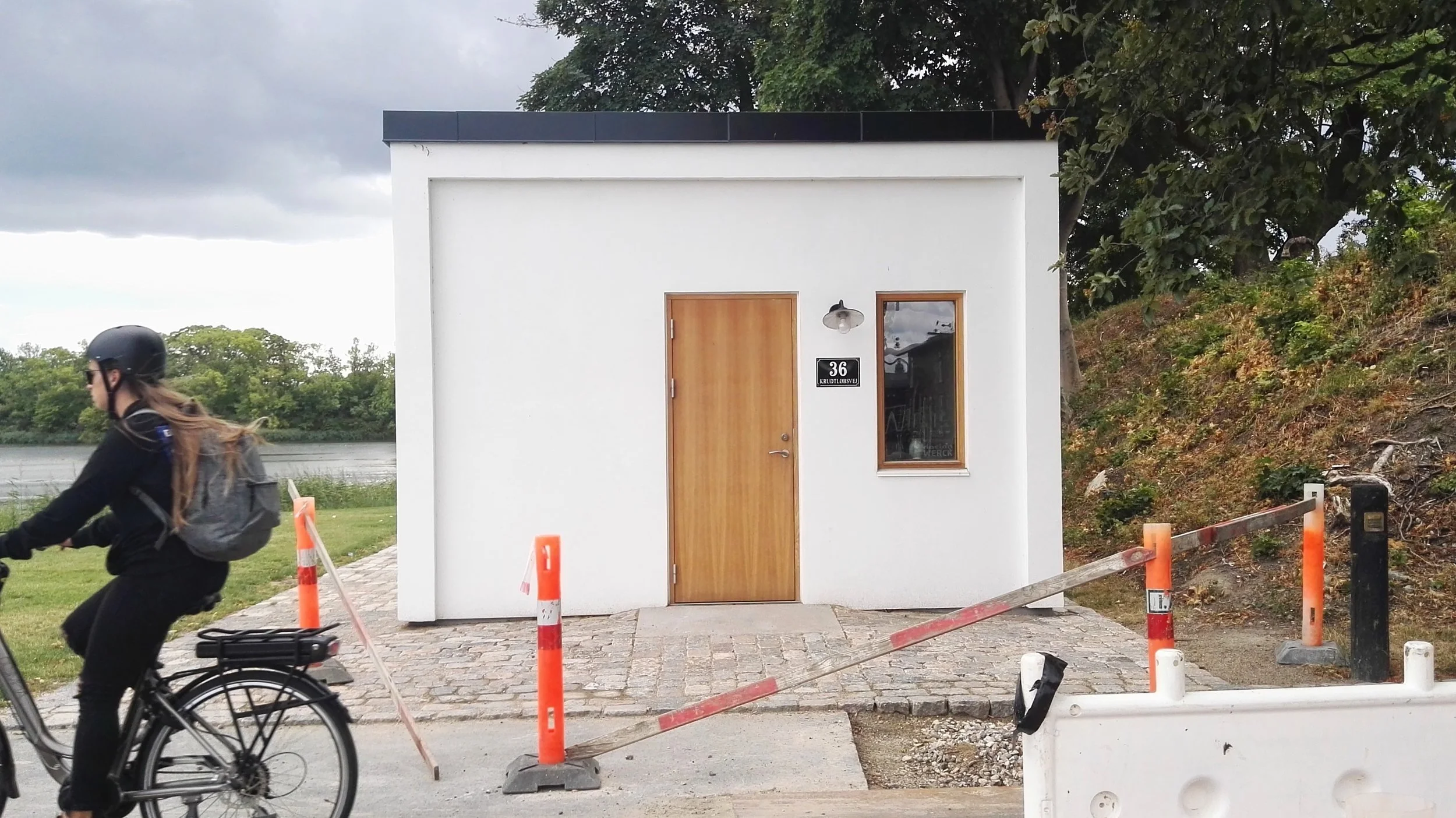 Casa moderna de color blanco con puerta de madera y una ventana, en una calle con andén de adoquines y señalización para construcción. En primer plano, una mujer montando bicicleta pasa frente a la casa, usando casco y mochila. Copenhagen.