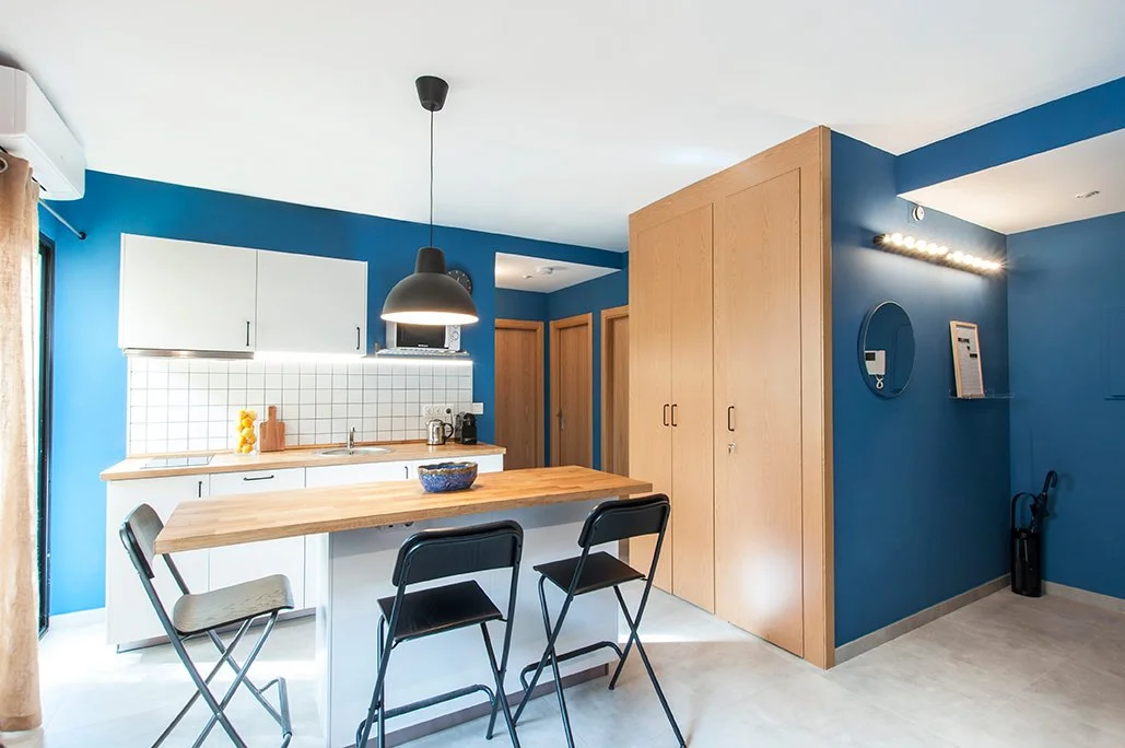 Cocina moderna con paredes azules, muebles blancos, una mesa de madera clara con tres sillas negras y una lámpara colgante negra.