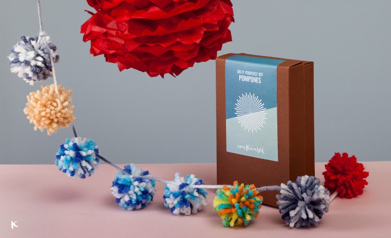 Una caja de bricolaje para hacer pompones, decoraciones de papel en color rojo y pompones de lana de varios colores en un fondo de pared gris y superficie rosa.