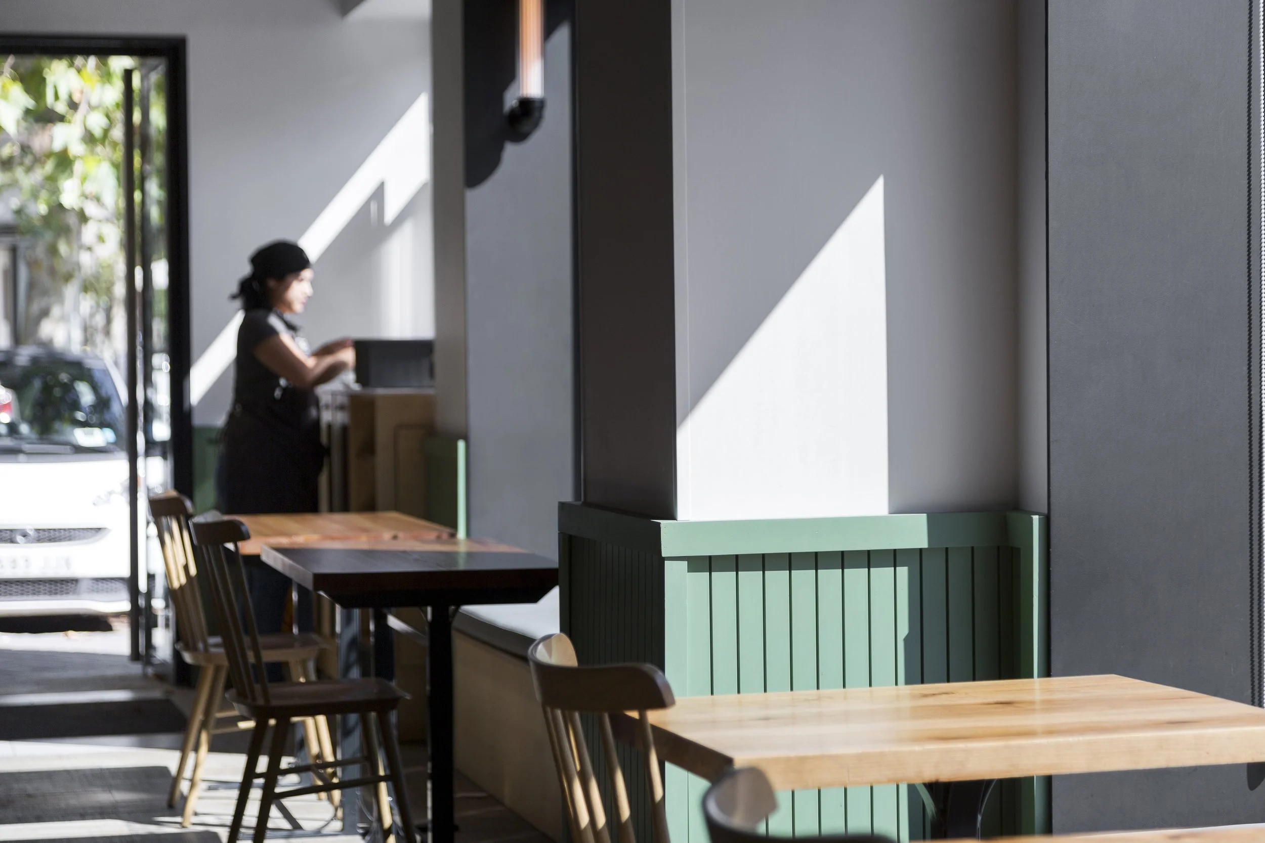 Interior de una cafetería con mesas de madera, sillas, y una persona de pie cerca de una pared iluminada por luz natural.