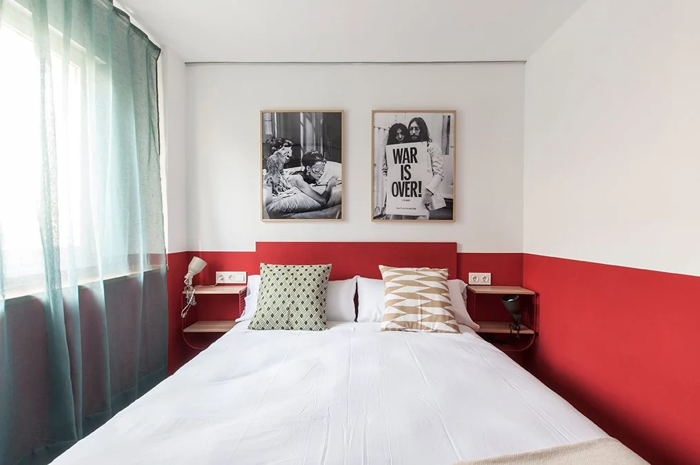 Habitación con cama blanca, cojines decorativos, paredes en tonos rojos y blancos, cuadros en la pared, y cortina verde.