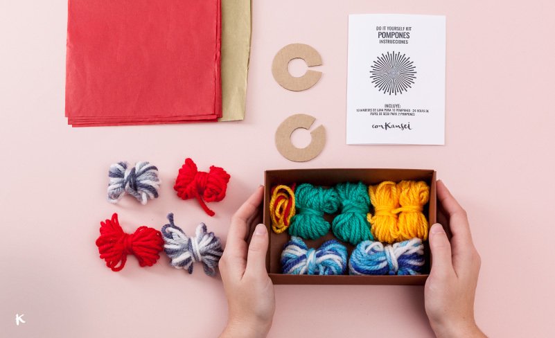 Manos sosteniendo una caja con ovillos de hilo de colores, junto a pompones de lana en tonos rojos, azules y verdes, y otros materiales para hacer pompones, en una mesa de fondo rosa.