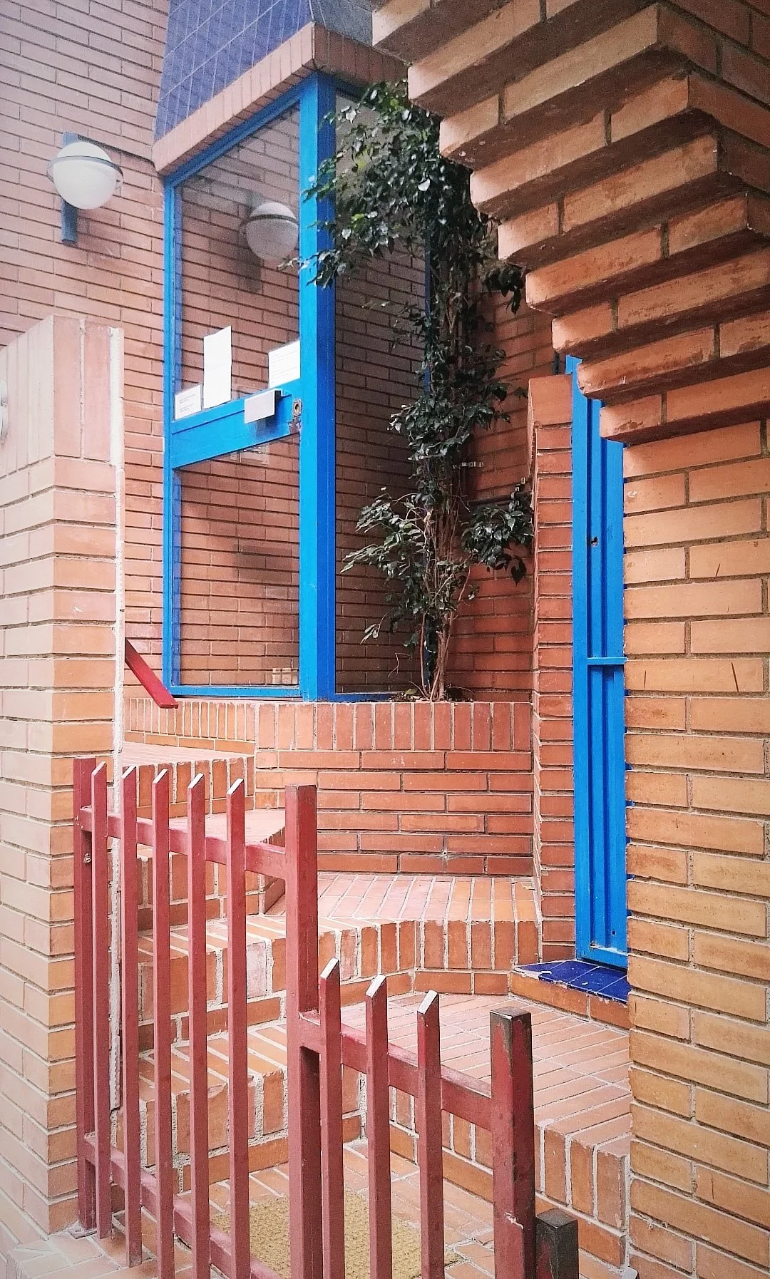Entrada de edificio con paredes de ladrillo, barandilla de madera pintada de rojo y estructura de marco azul, con planta trepadora y ventanas con cristalería moderna.