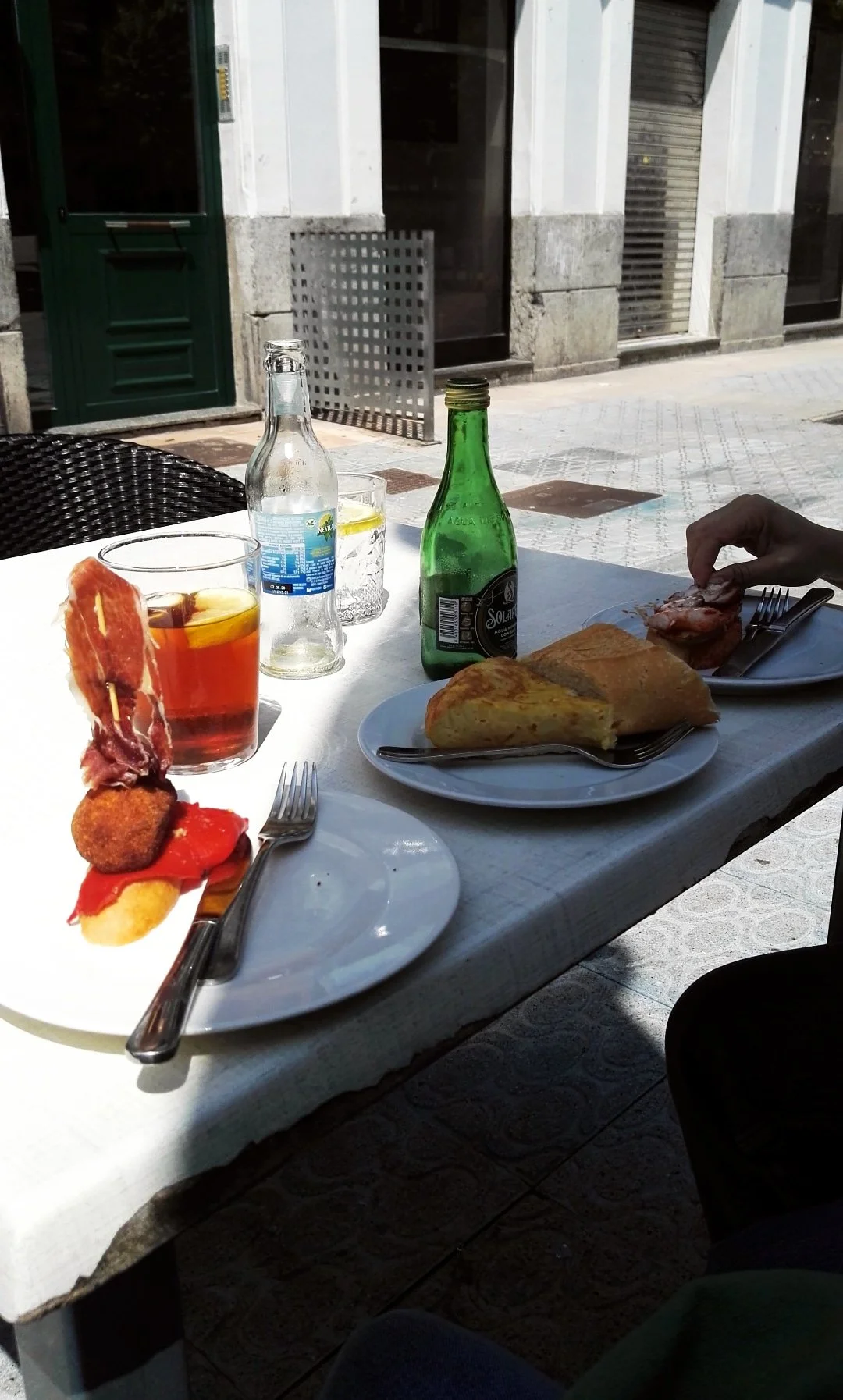 Mesa con comida y bebidas, incluyendo una rebanada de tortilla española, una porción de jamón, una empanada y una ensalada, en un restaurante en la calle.
