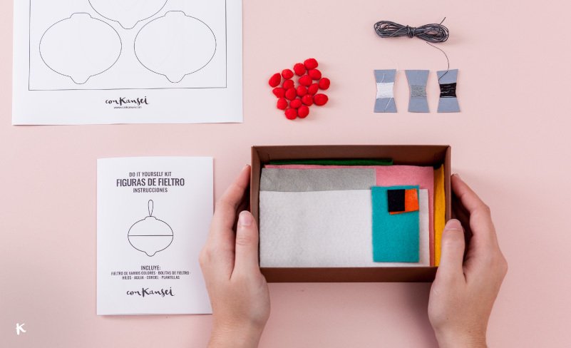 Manos sosteniendo una caja con materiales de manualidades para hacer figuras de filtro. Hay bolas rojas, hilos, papel y un instructivo. También se ven moldes, fichas de colores, y un diagrama de una figura en una hoja en un fondo rosa claro.