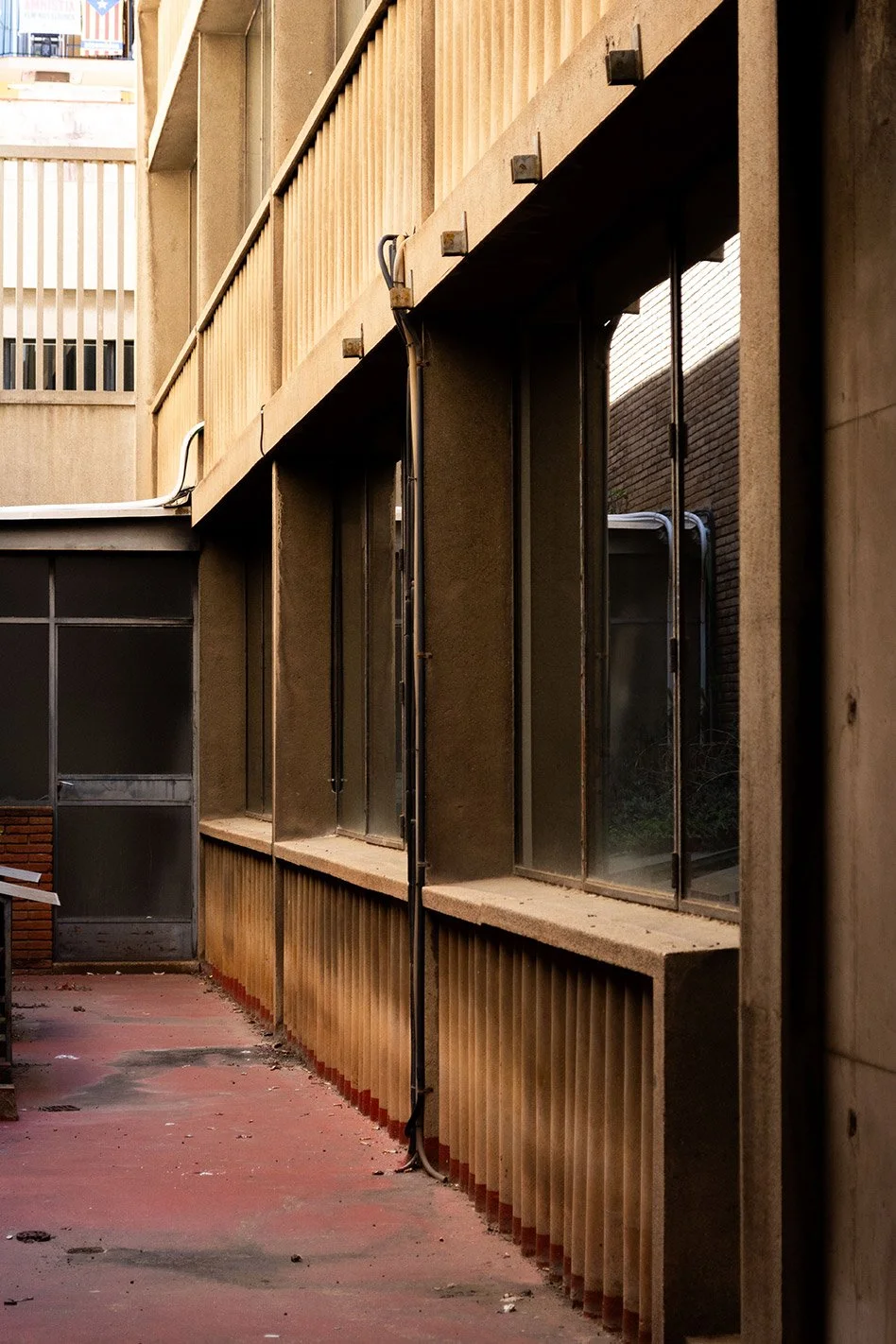 Vista de una fachada de edificio con ventanas de vidrio y barandilla de madera, en un pasillo exterior con piso de concreto.