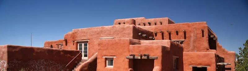 Casa de estilo adobe bajo un cielo azul, con estructura de arcilla roja y vigas de madera sobresalientes.
