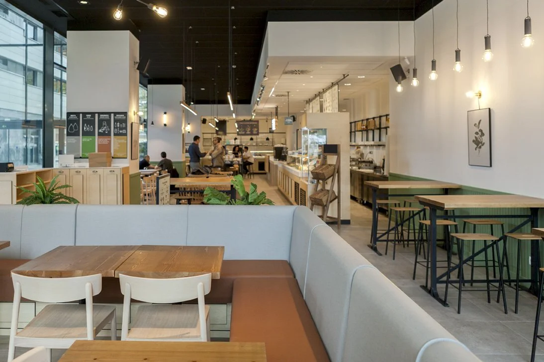 Interior de una cafetería moderna con mesas y sillas de madera, iluminación colgante y paredes blancas. Al fondo, hay personas sentadas y un mostrador de servicio con vitrinas. Diseño de interiorismo comercial. Retail Design.