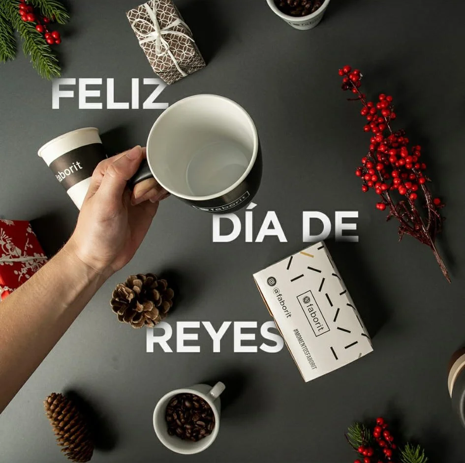 Taza siendo sostenida por una mano, rodeada de objetos navideños como un regalo envuelto, piñas, ramas con frutos rojos y una caja de café. Texto "Feliz Día de Reyes". Diseño redes sociales. Design for social media. Agosto Creative Studio, Barcelona.