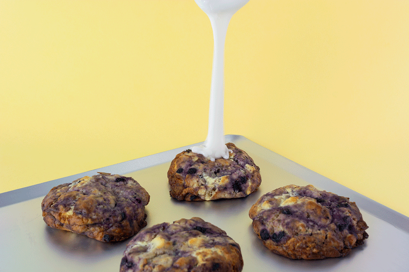 Vasos de galletas con chispas de chocolate con leche y avena, con leche siendo vertida sobre una de ellas en un fondo de paredes amarillas. Mionas Bakery. Especialidad en Cookies en Sitges. Retail Design, diseño de tiendas. Foto de producto.