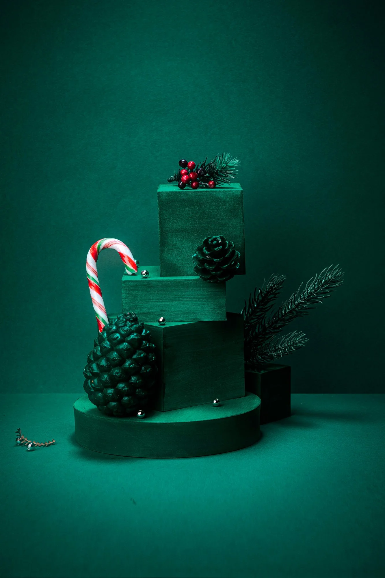 Decoración navideña con piñas, bastón de caramelo, ramas de pino, bolas plateadas y bloques de madera en tonos verdes sobre fondo verde oscuro.