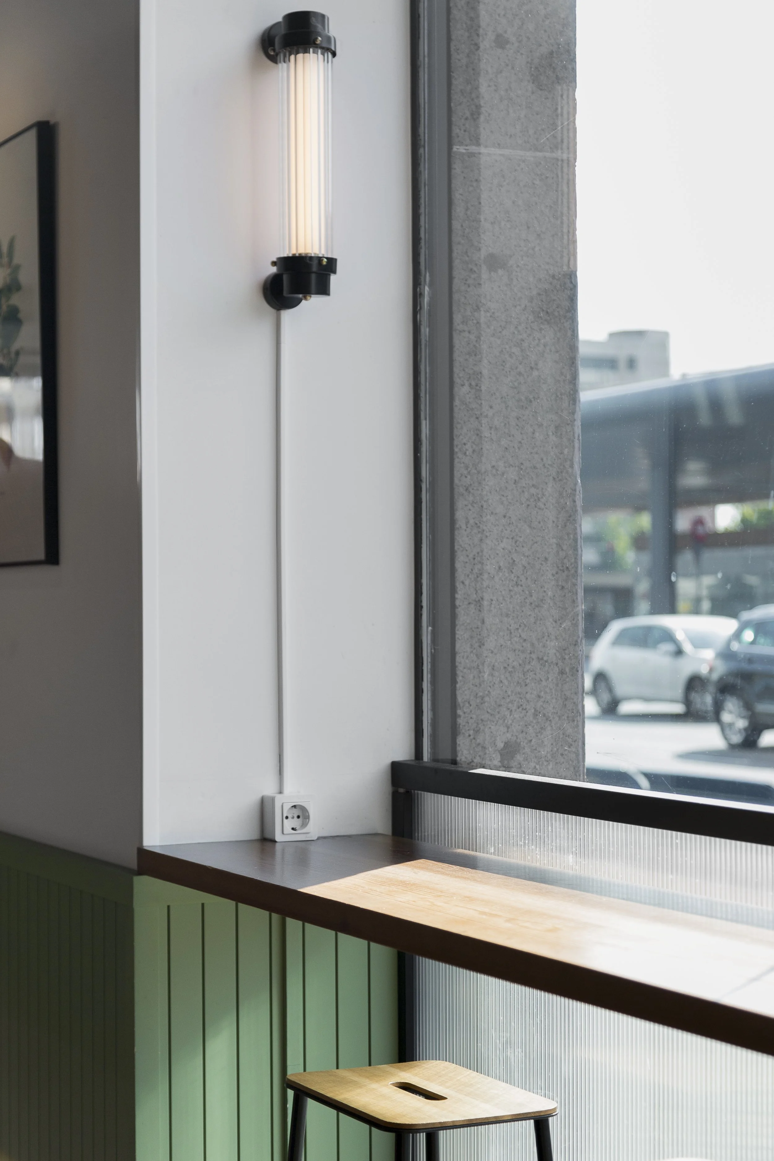 Mesa de madera con taburete en una cafetería junto a una ventana, con una lámpara de pared y un tomacorriente visible. Diseño de interiorismo comercial. Retail Design.