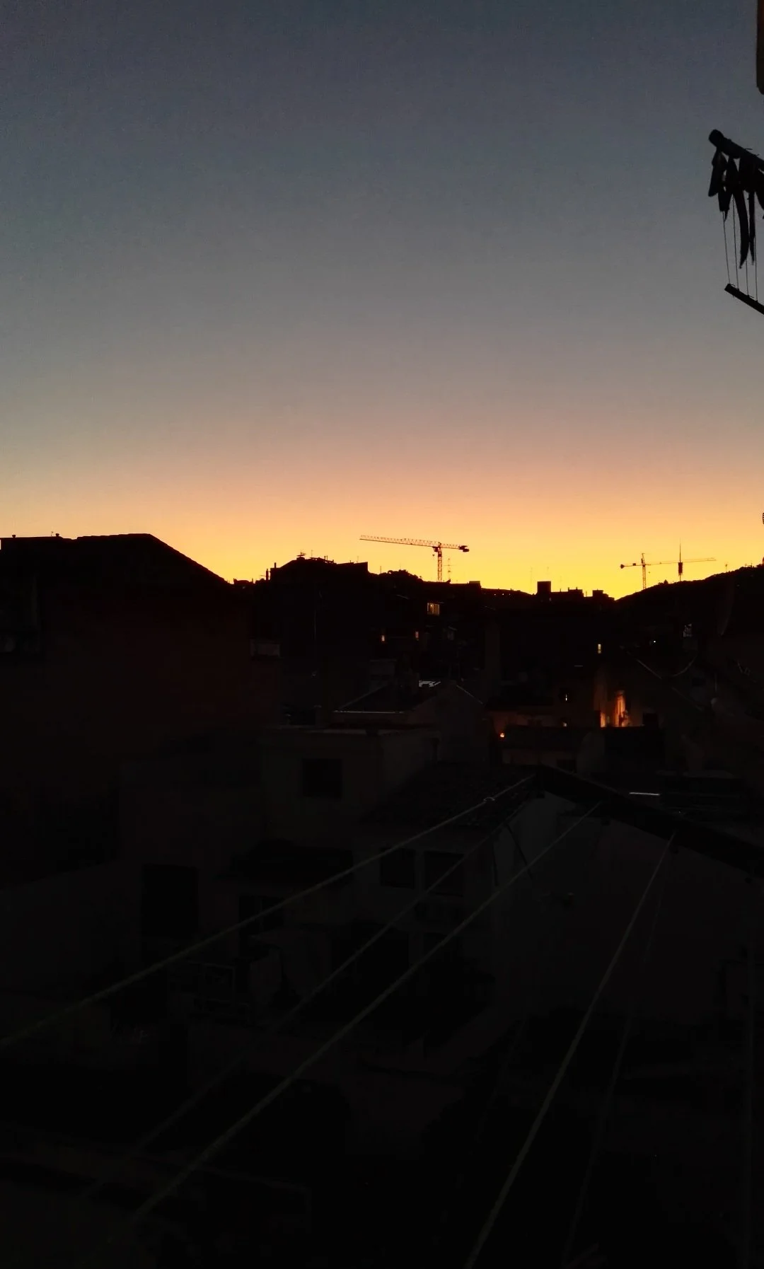 Atardecer en una ciudad con siluetas de edificios y grúas de construcción, cielo con tonos oscuros y una línea luminosa en el horizonte.