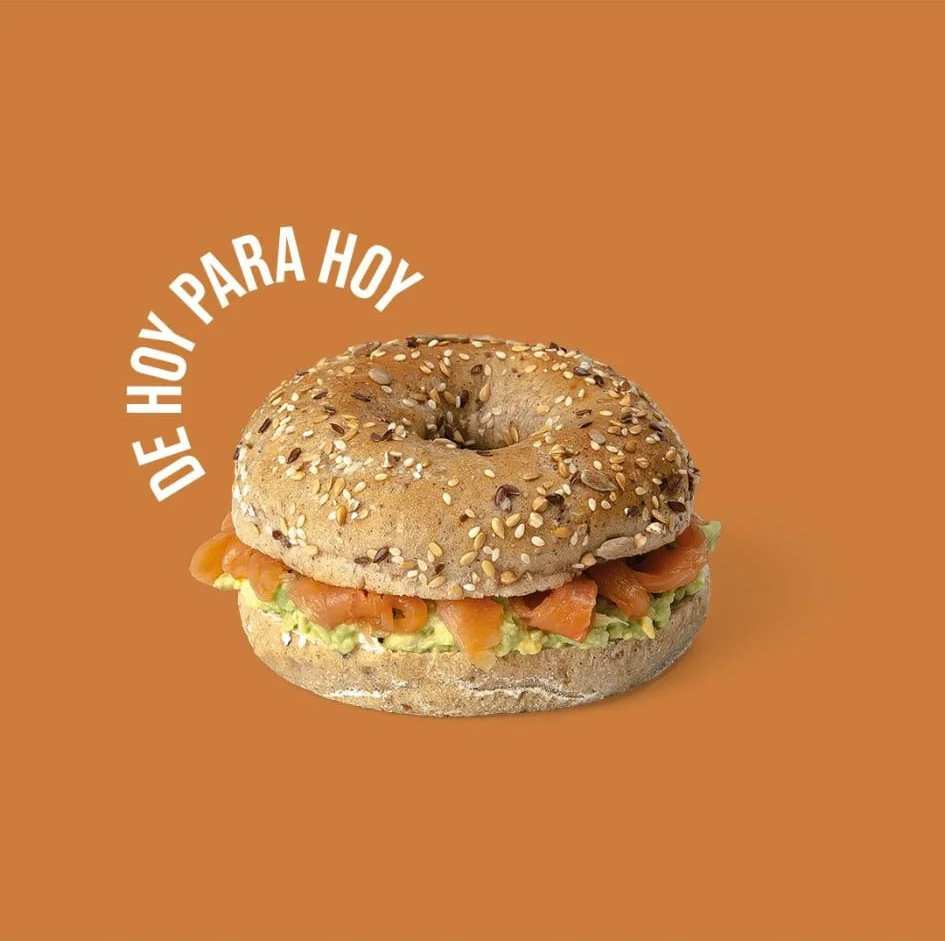 Bagel de semillas relleno de salmón y aguacate sobre fondo naranja con texto "De hoy para hoy". Diseño de redes sociales.  Design for social media. Agosto Creative Studio, Barcelona.