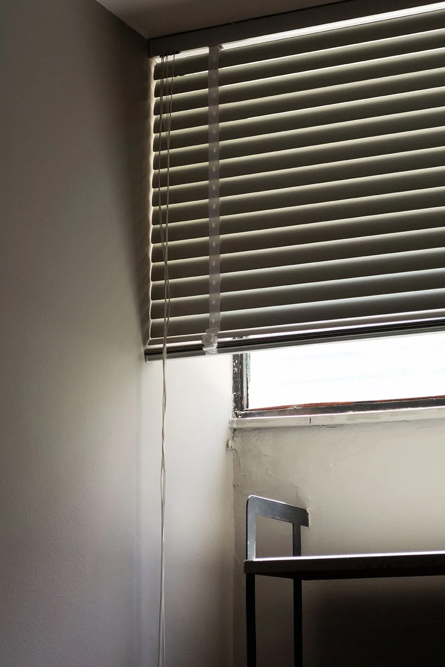 Ventana con persianas horizontales cerradas en una habitación con pared blanca y una silla de metal al lado.