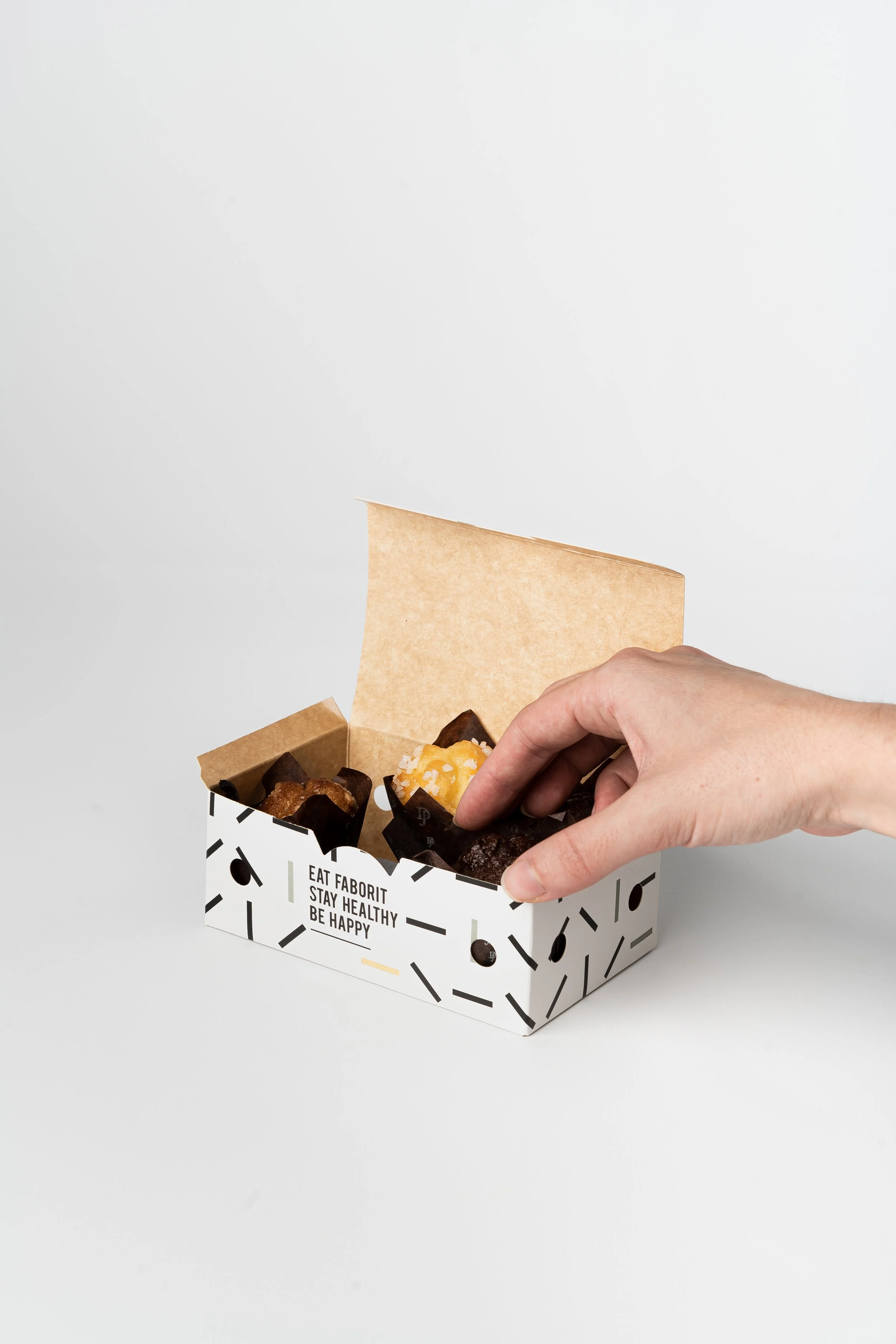 Caja abierta de cartón con diseño blanco y negro, contiene alimentos y muestra una mano tomando uno de ellos.