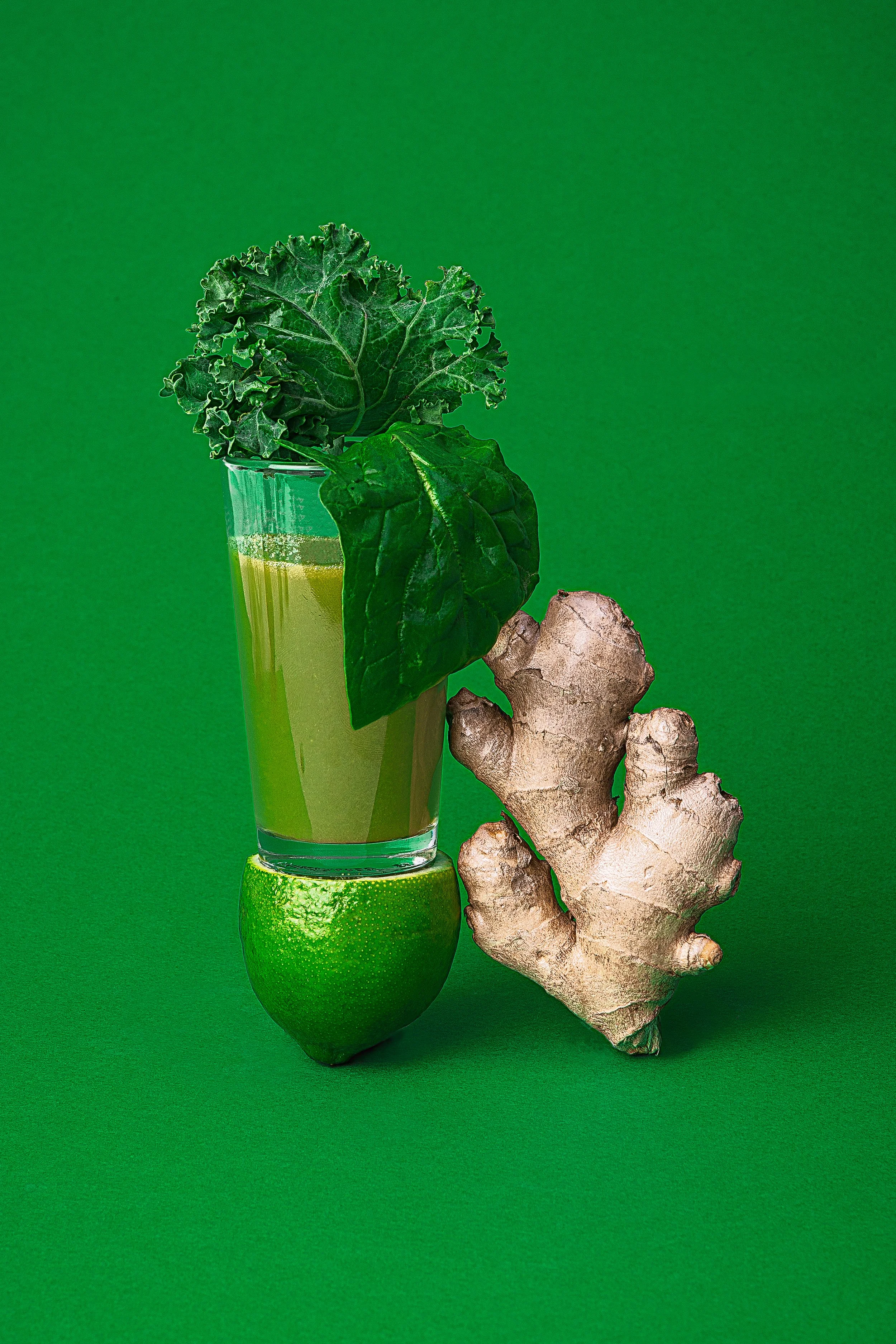 Un vaso con jugo verde con hojas de kale y perejil, una lima, jengibre y medio limón sobre un fondo verde.