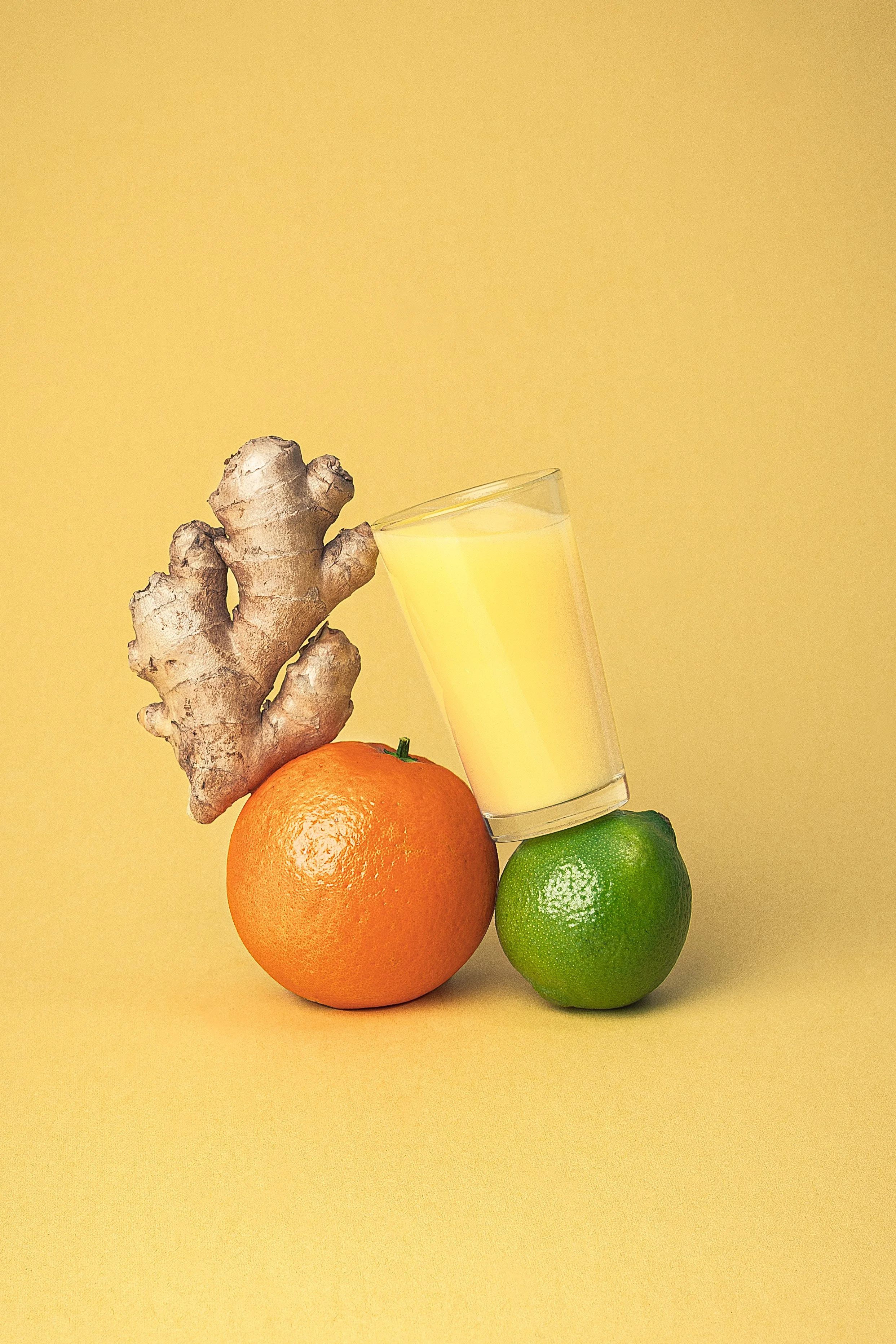 Jengibre, naranja, limón y un vaso con jugo de limón sobre un fondo amarillo.
