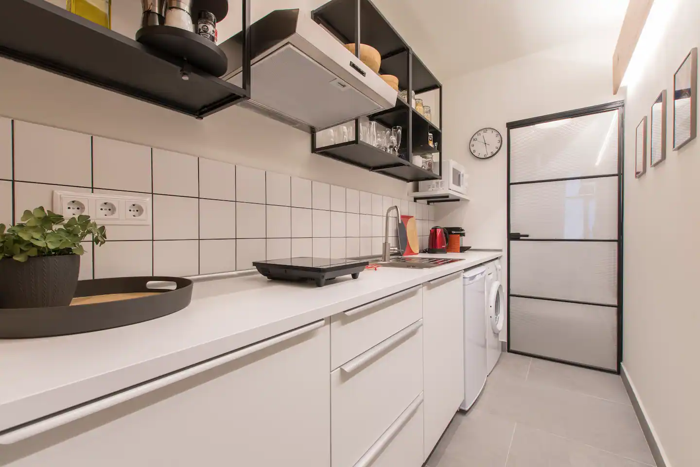 Cocina moderna y minimalista con encimera blanca, armarios blancos, estantes abiertos en color negro, utensilios de cocina y electrodomésticos, como cafetera y microondas, y una puerta de madera con paneles de vidrio.