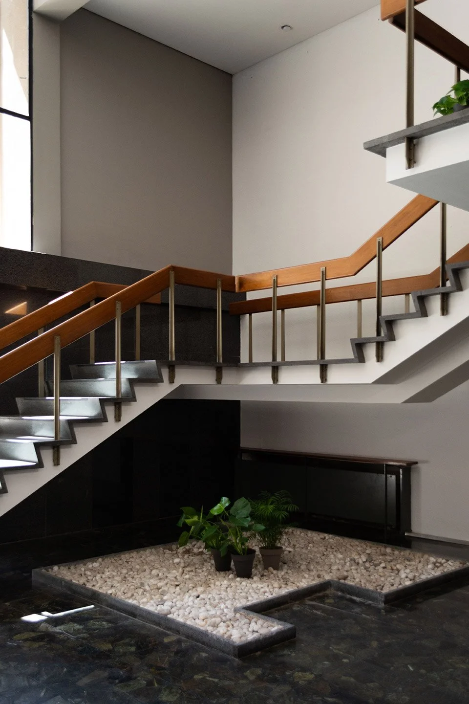 Escalera interior de edificio moderno con barandales de madera y pasamanos de metal, área decorada con plantas y grava blanca.