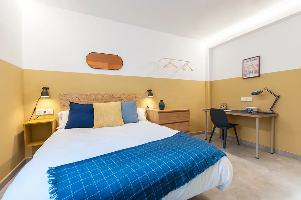 Habitación moderna con cama doble, edredón blanco, cojines de colores, alfombra azul y escritorio con silla negra. Decoración minimalista y pared bicolor.