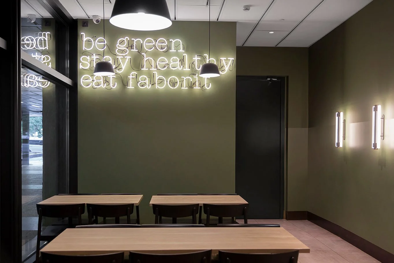 Interior de un restaurante moderno con paredes verdes, mesas de madera, lámparas colgantes negras y un letrero de neón que dice "be green stay healthy eat fab".