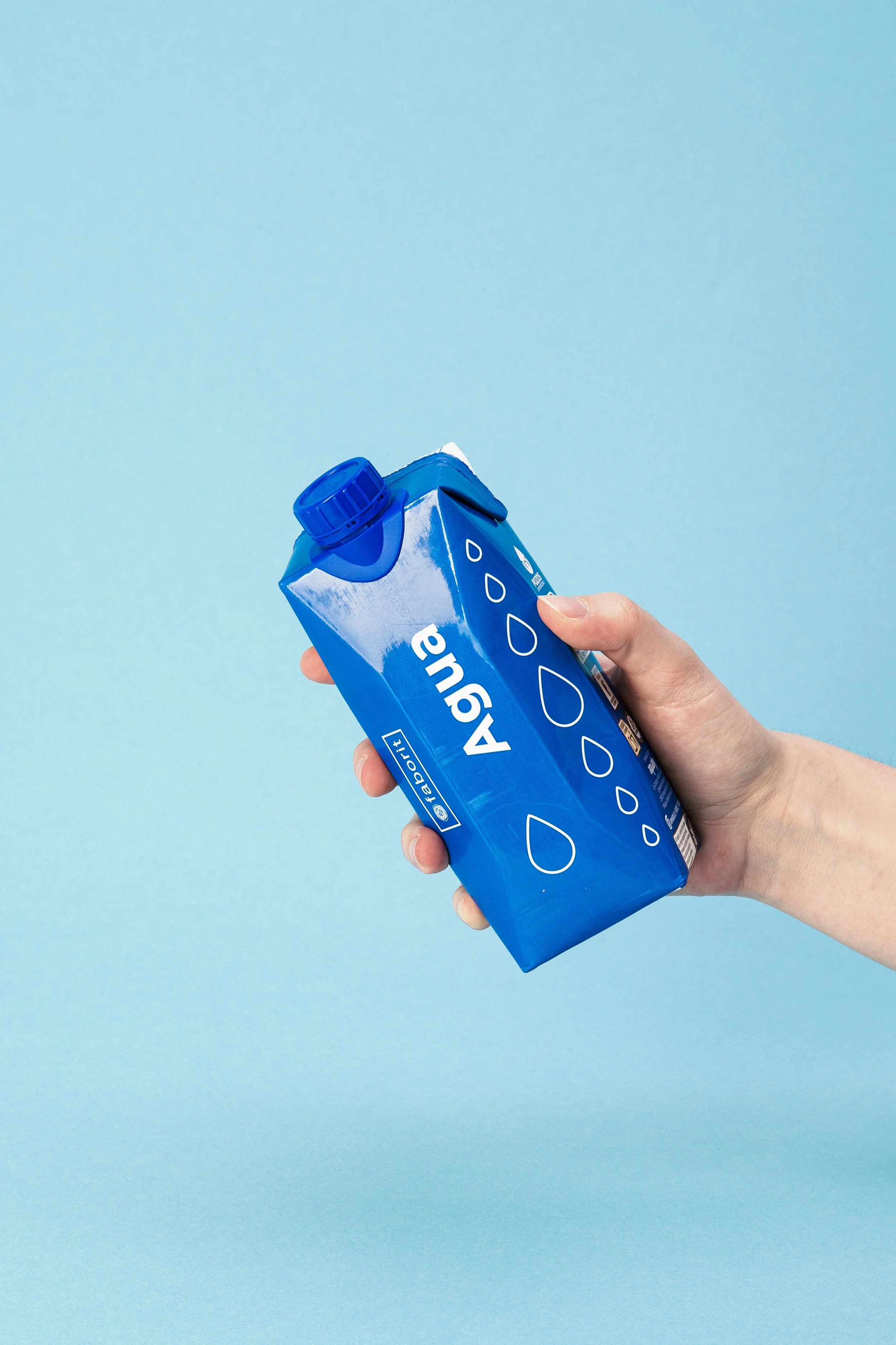 Mano sosteniendo un cartón de agua azul sobre un fondo azul claro. Diseño de packaging. Agosto Creative Studio.