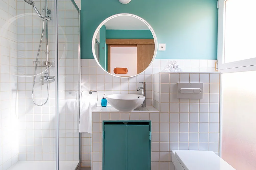 Baño moderno con ducha de vidrio, lavabo con espejo redondo, toallas blancas, y decorado en tonos turquesa y blanco.
