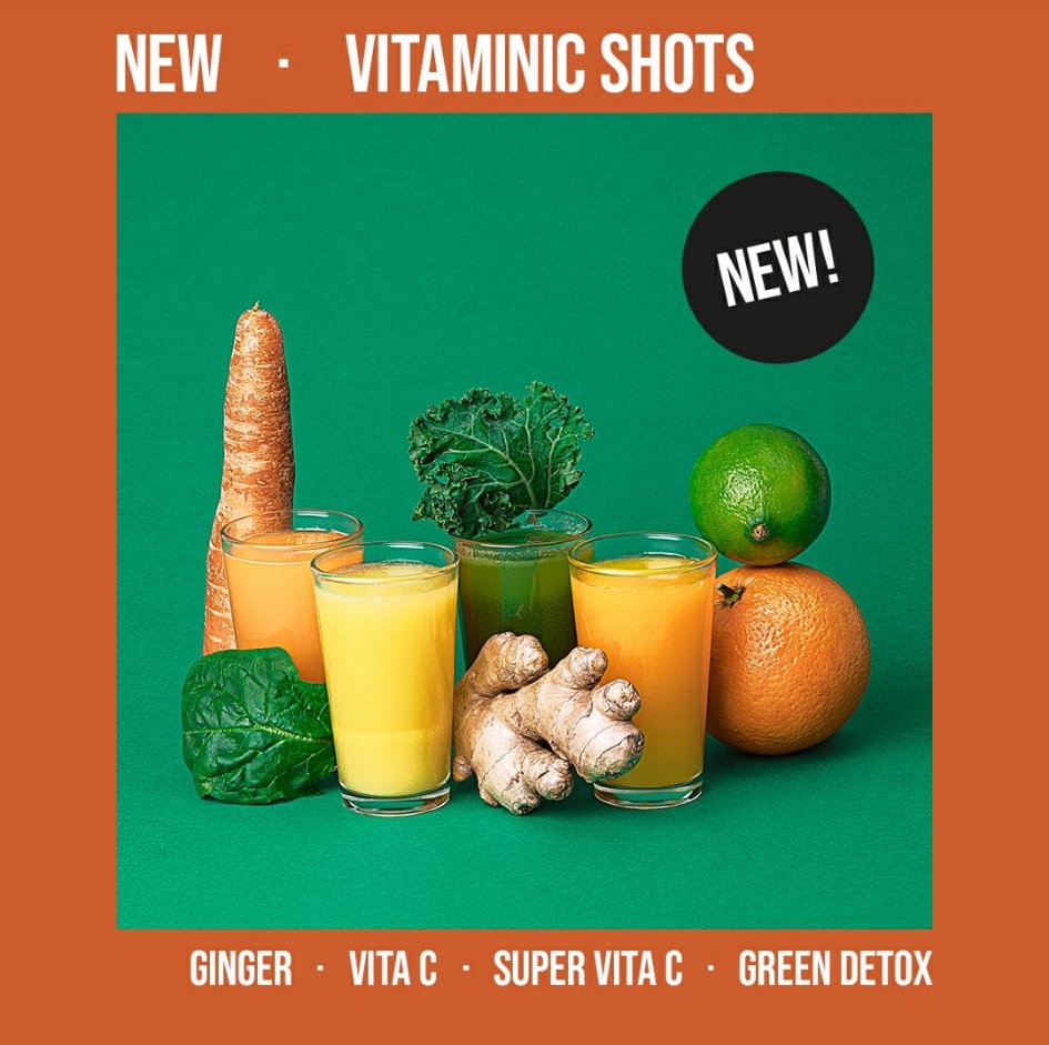 Imagen de cuatro vasos con jugos de diferentes colores acompañados de ingredientes frescos como jengibre, zanahoria, hojas verdes, naranja y limón sobre un fondo verde. Texto promociona "Vitaminic Shots" nuevos con sabores: Jengibre, Vitamina C, Súpe