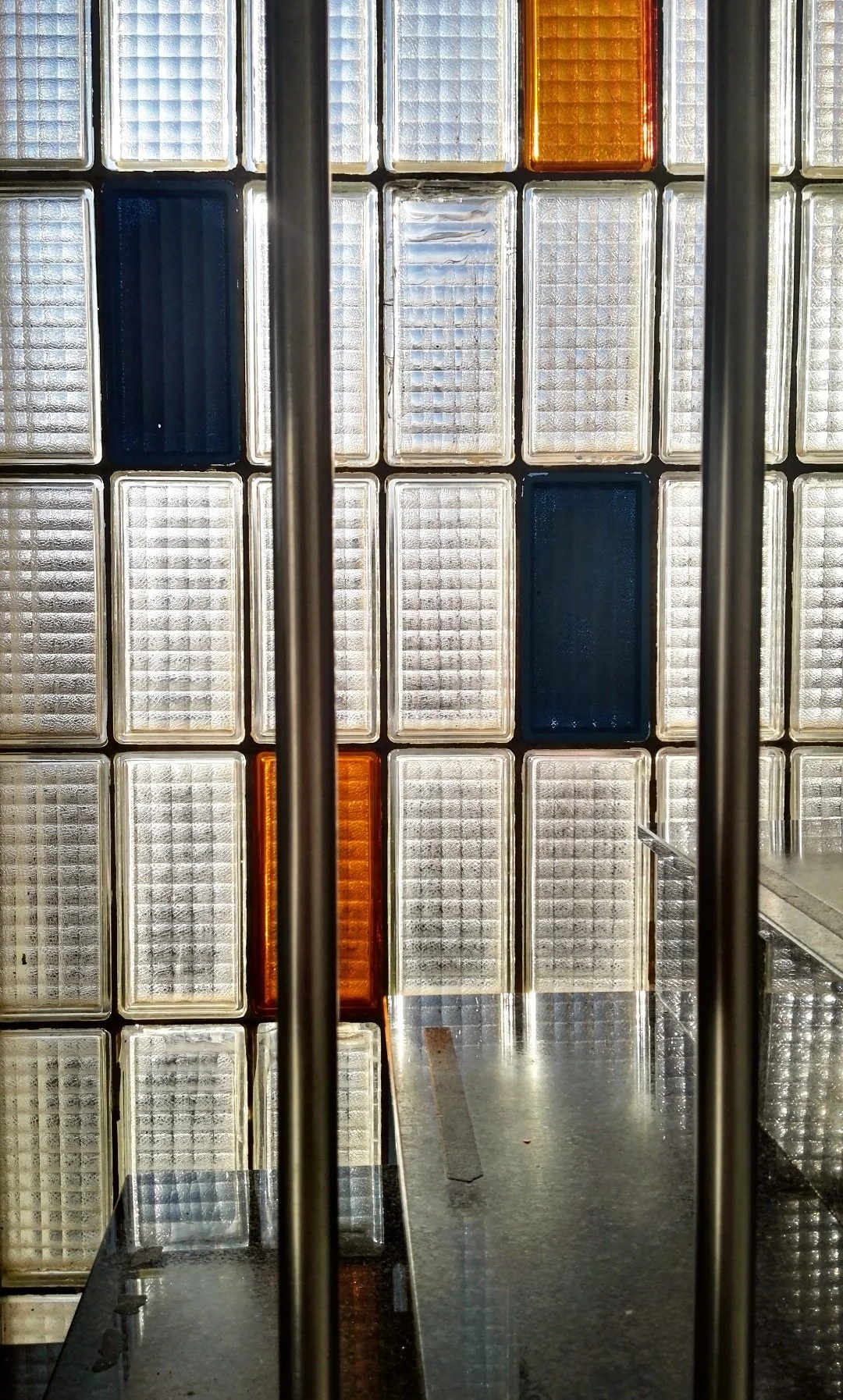 Ventanas de vidrio con bloques de diferentes colores, entre ellos blanco, azul, y naranja, vista a través de una estructura metálica y una mesa de piedra o mármol.