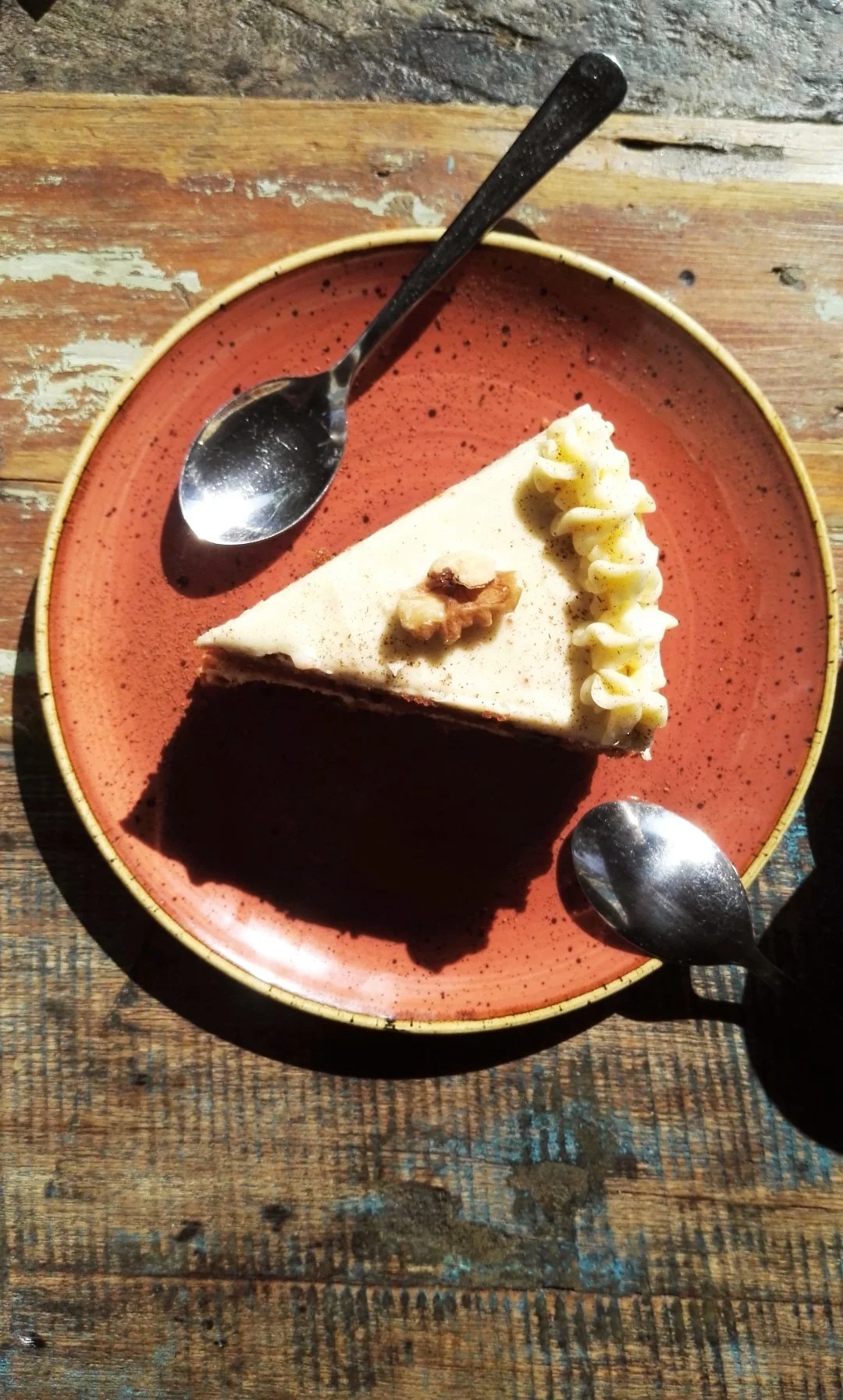 Porción de pastel de chocolate con glaseado claro, decorado con crema en forma de ondas y una nuez, servido en un plato naranja con dos cucharas. Fondo de madera rustic.