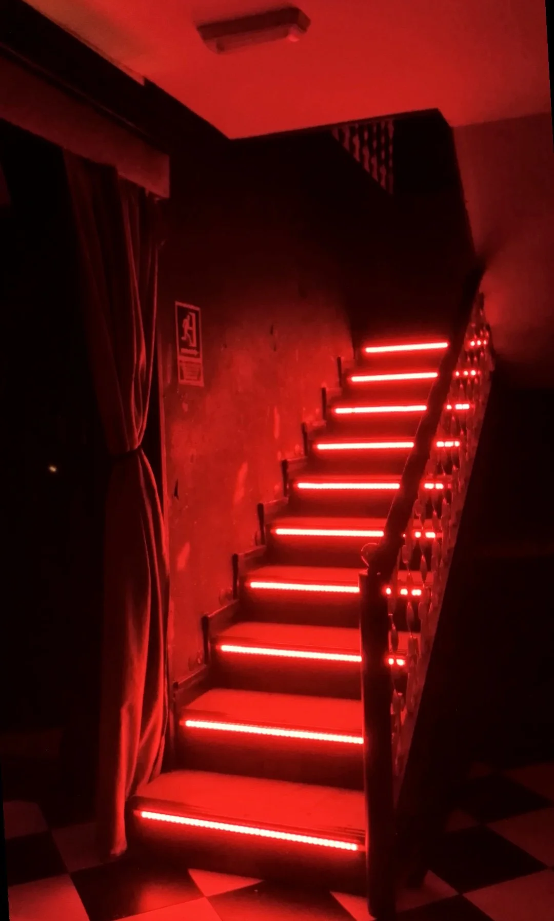 Escalera con luces rojas en un ambiente oscuro, con suelo de baldosas negras y blancas y cortinas rojas a un lado.