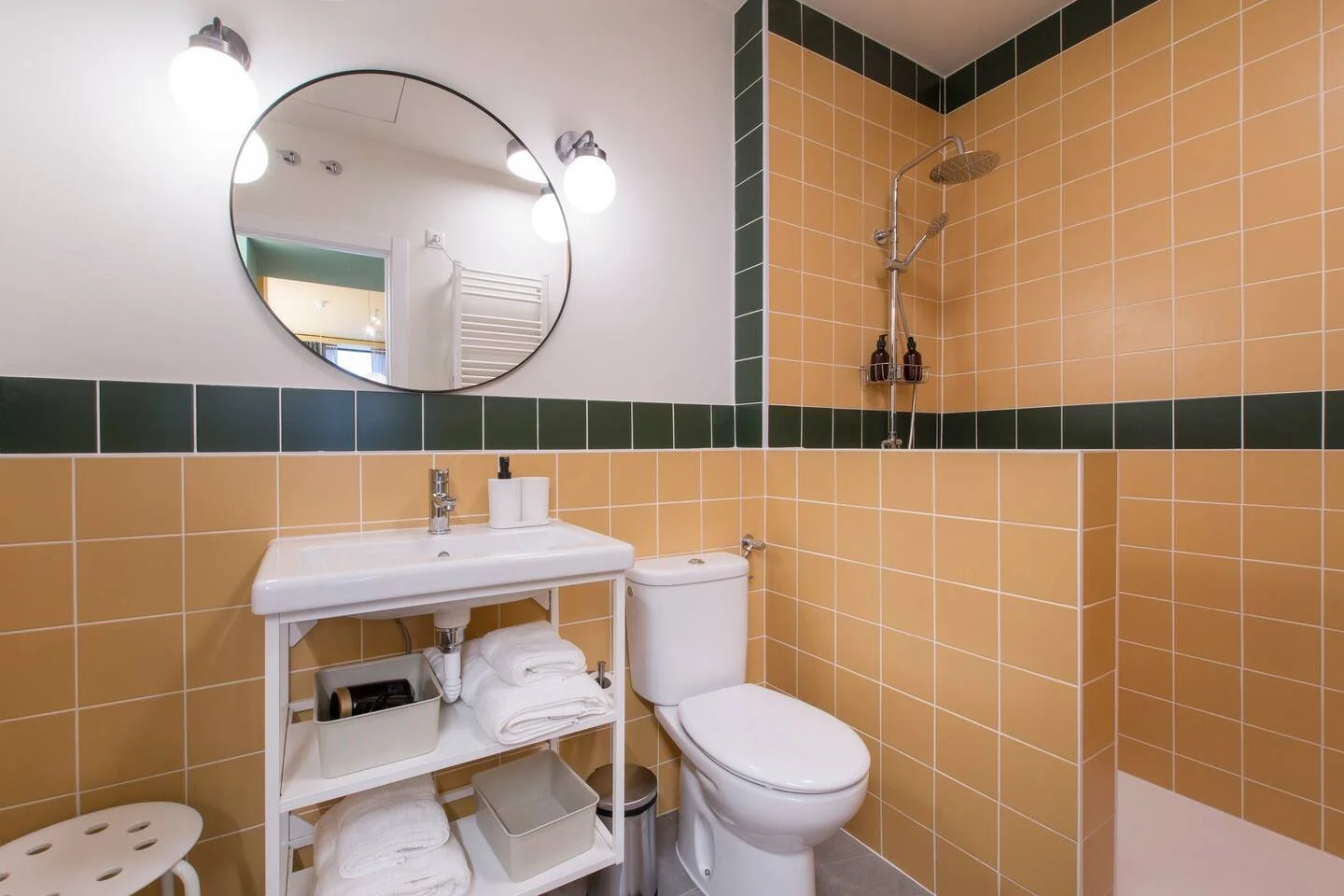 Baño moderno con paredes de azulejo en tonos beige y verde, espejo redondo, lavabo con estantería, inodoro y ducha con cabecera fija.