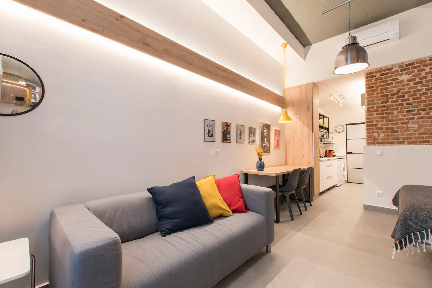 Sala de estar moderna con sofá gris, cojines en colores negro, amarillo y rojo, mesa con sillas, decoración de cuadros en la pared, y área de cocina con electrodomésticos y utensilios.