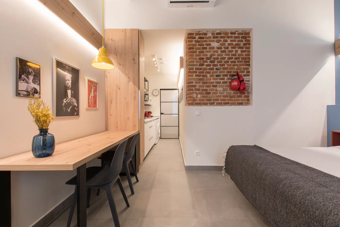 Interior de un apartamento con pared de ladrillos expuestos, cama, escritorio con sillas, decoraciones y cocina en el fondo.