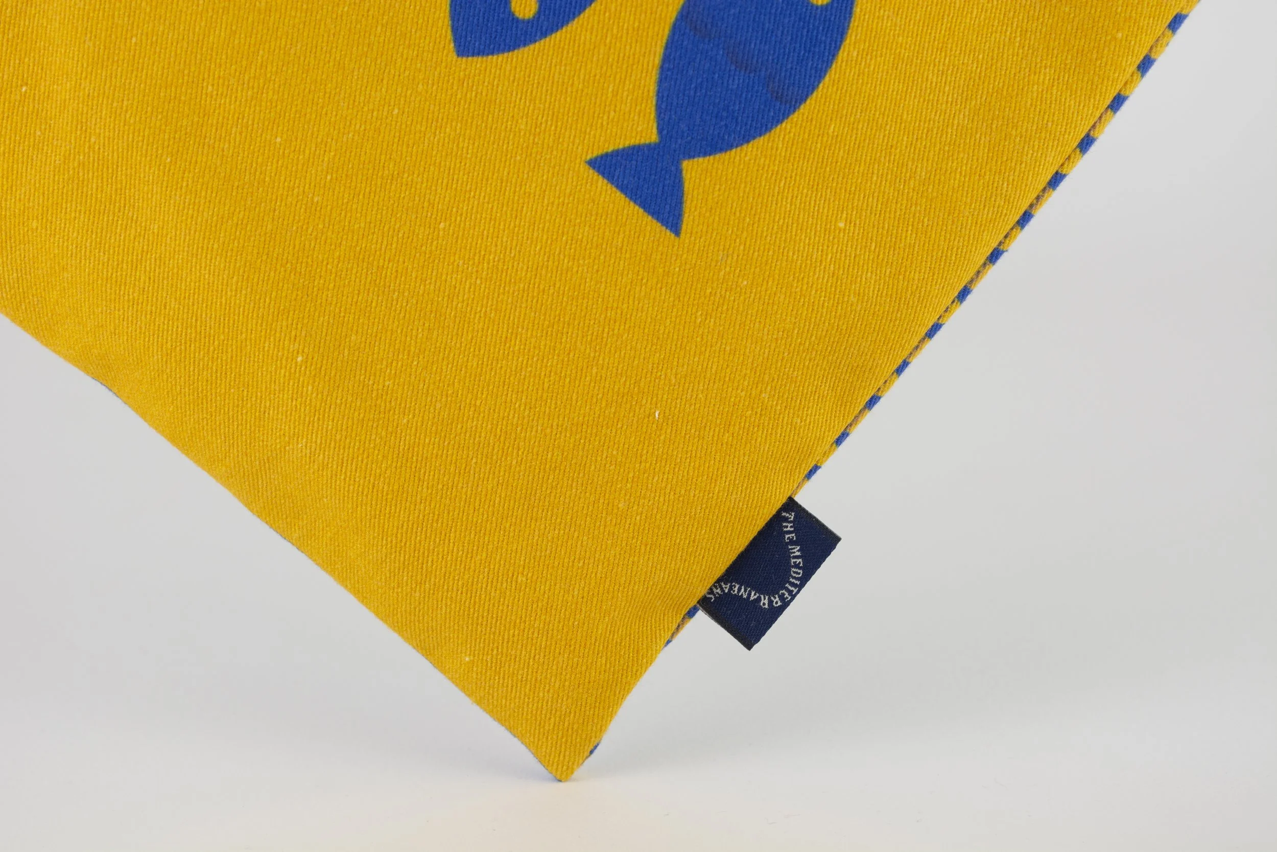 Imagen de una esquina de un cojín amarillo con un diseño de pez azul, y una etiqueta azul en la esquina inferior derecha.