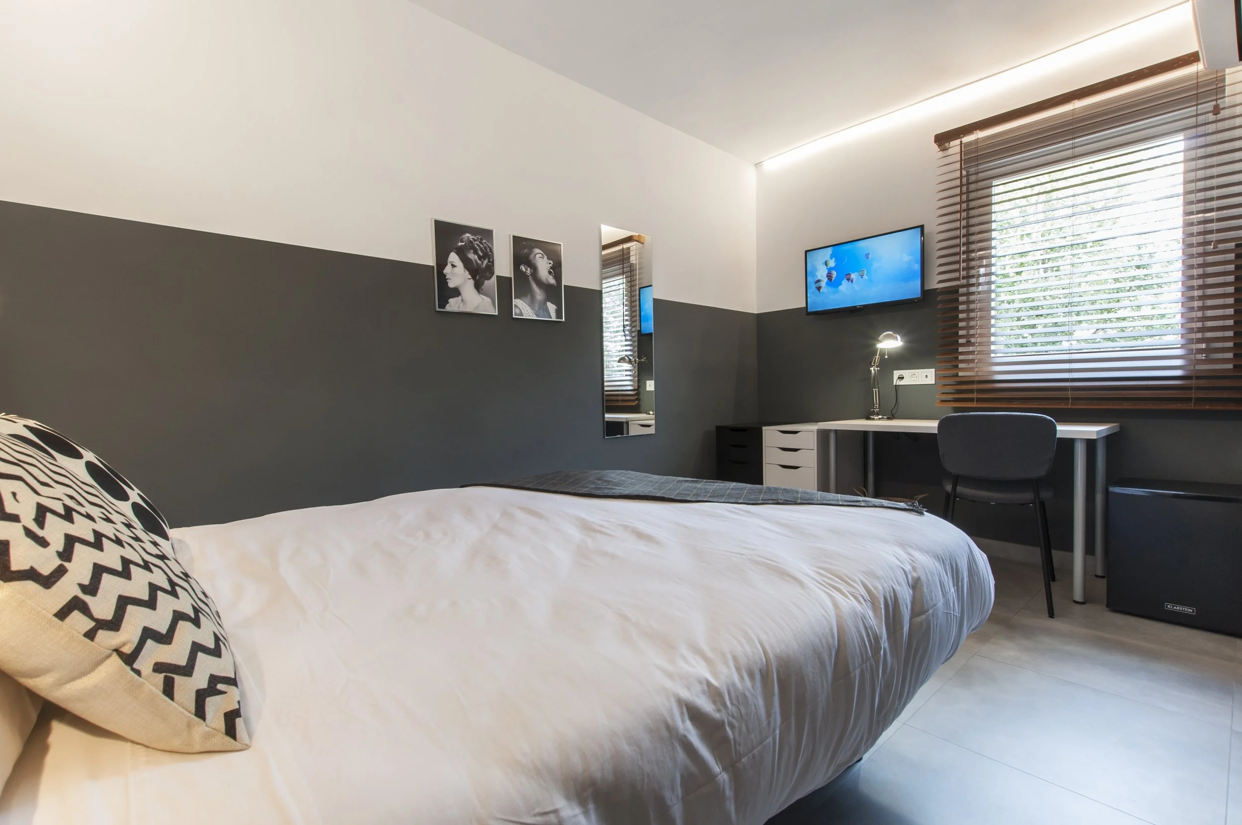Habitación moderna con cama, escritorio, televisor, ventanas con persianas y cuadros en la pared.