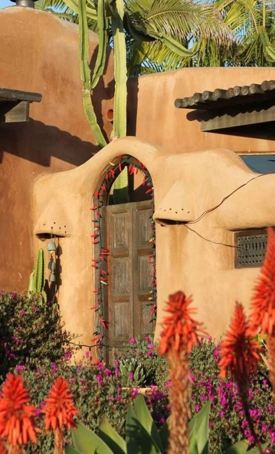 Casa de adobe estilo suroeste con una puerta de madera rodeada de luces, cactus y plantas con flores naranjas y púrpuras.
