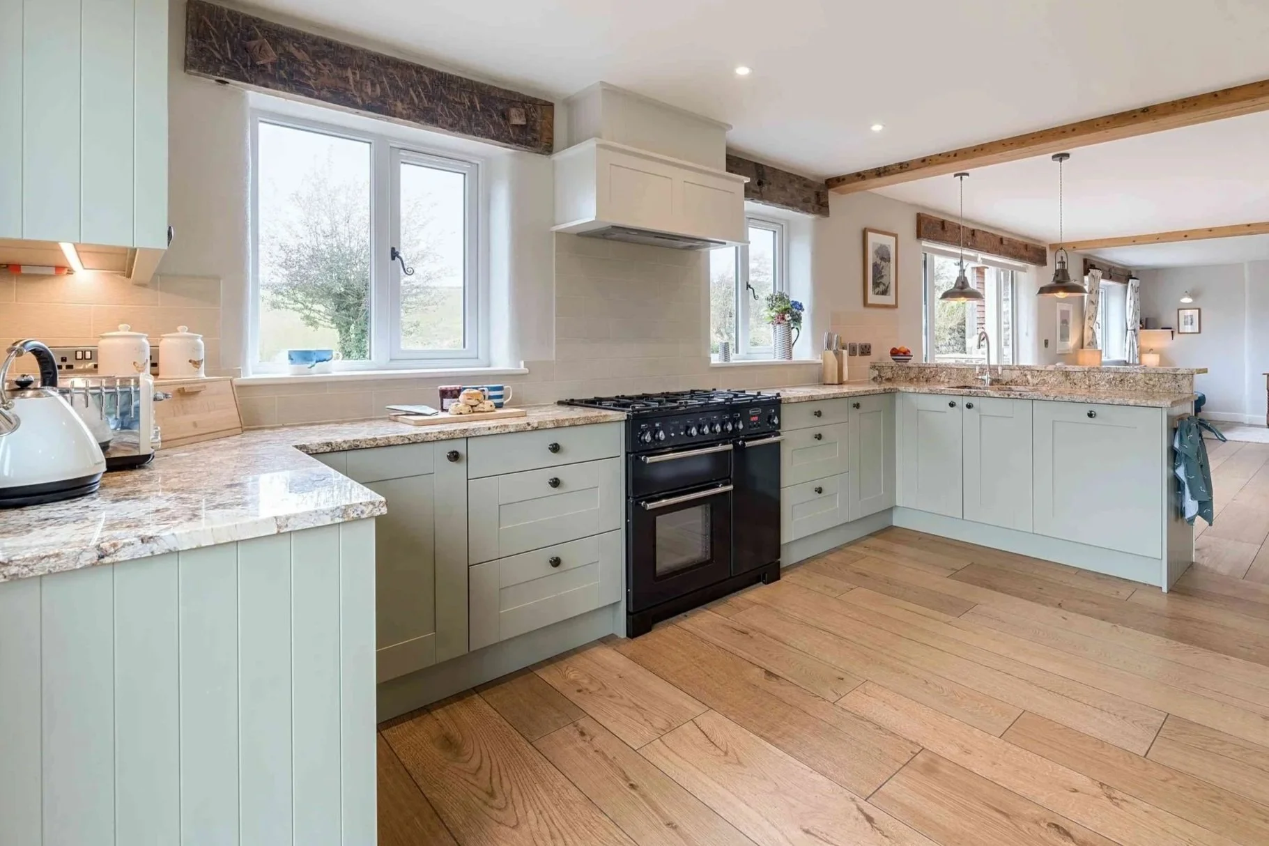 Open kitchen with light green cabinets, marble countertops, black stove, and white tiled backsplash, overlooking a dining and living area with wooden beams and windows.
